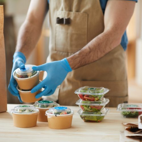 Closeup of unrecognizable worker wearing protective gloves packaging orders at wooden table in food delivery service, copy space